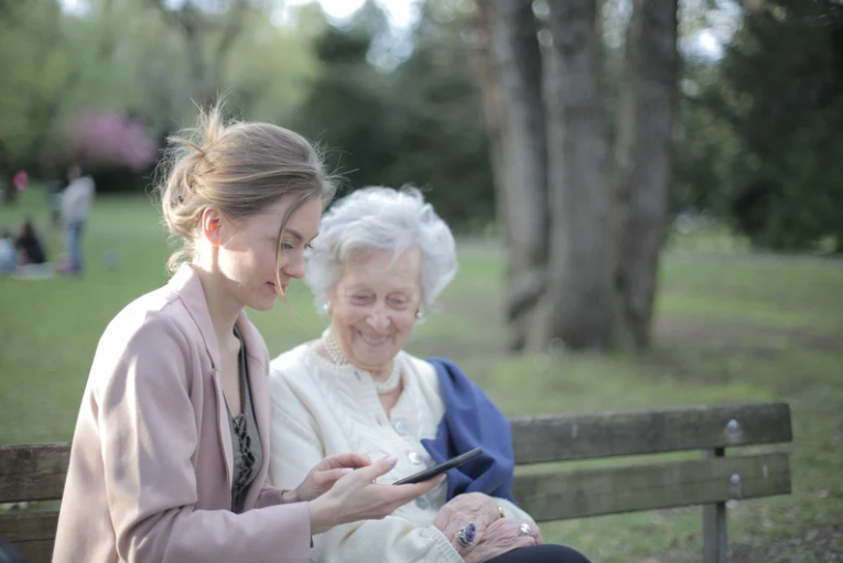A younger woman helping an elderly woman on a park bench and enjoying some media on a phone